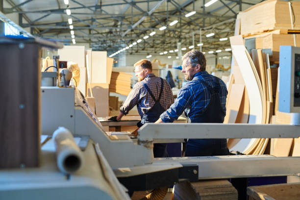 Serious confident industrial workers in overalls and safety goggles standing at workbenches and examining particleboard in assembly shop of furniture factory